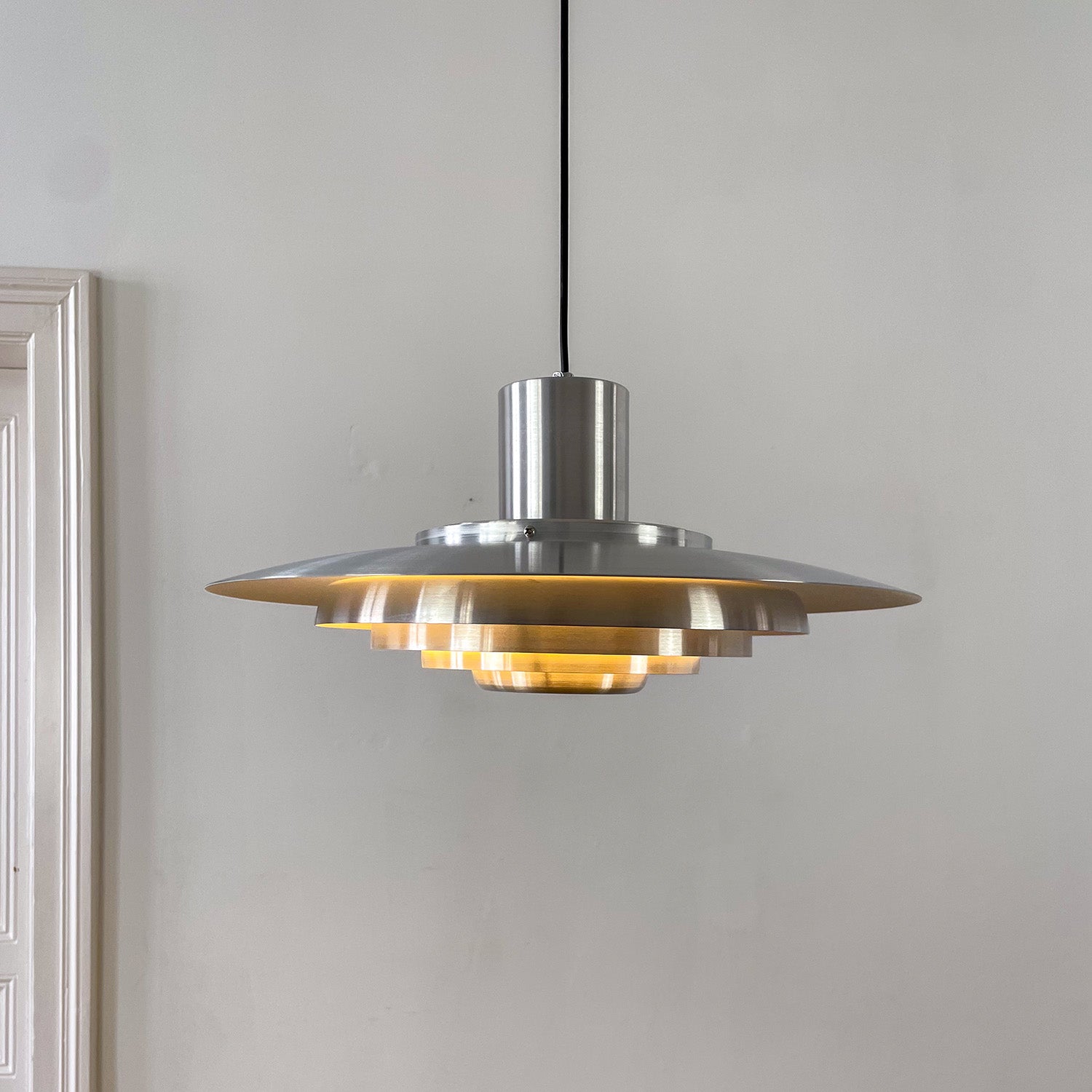 Chromlampe mit gestuftem Reflektorschirm, ideal für Esszimmer oder Büros. Der Hintergrund mit weißen Wänden und einer klassischen Altbau-Tür unterstreicht das reduzierte Design.