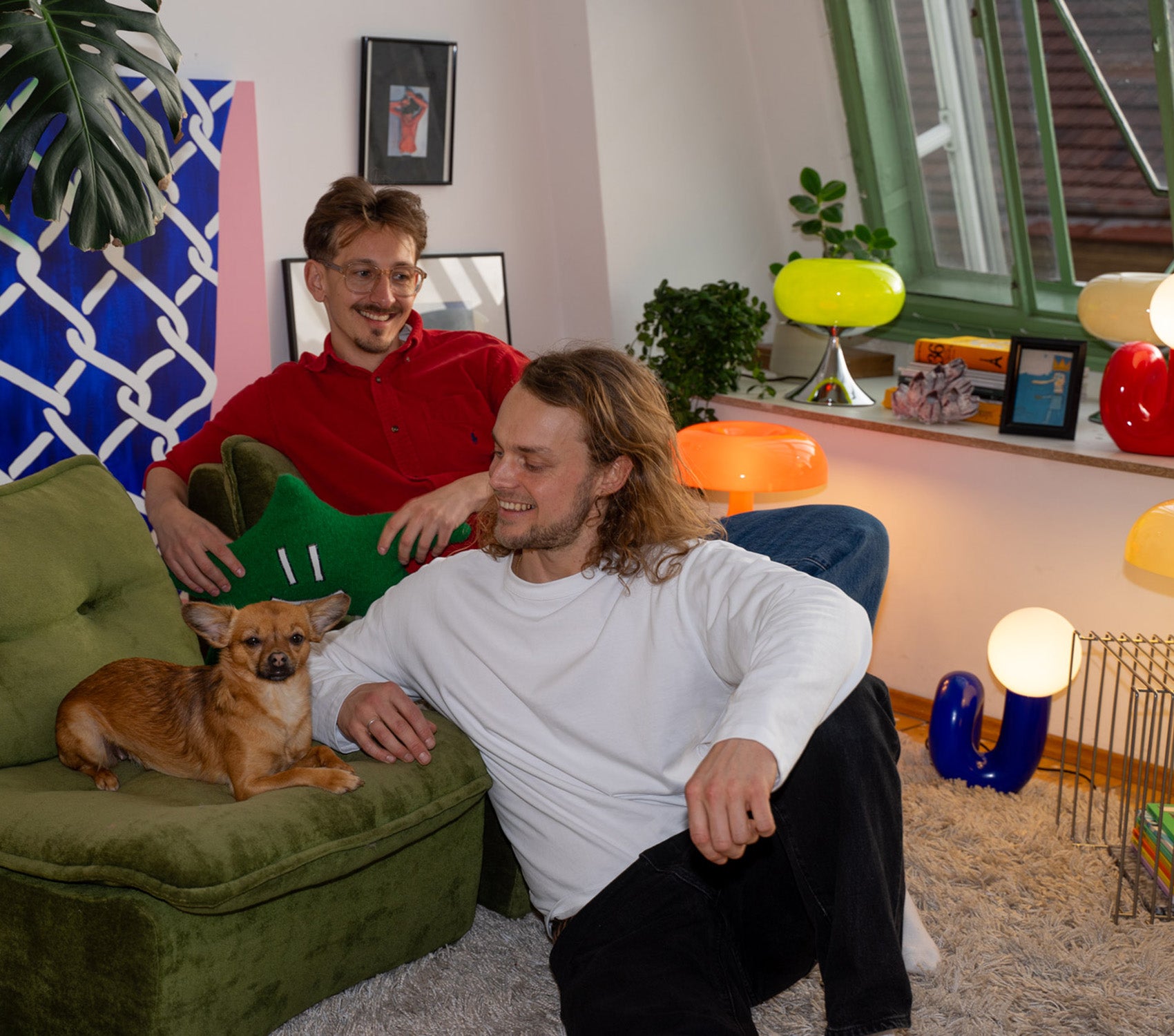 Gemütliche Szene im Wohnzimmer mit zwei Männern und kleinem braunen Hund auf grünem Retro-Sessel. Bunte Midcentury-Lampen, Pflanzen und Bilder auf der Fensterbank, Teppich und verspieltes Wandbild sorgen für eine warme, kreative Atmosphäre.