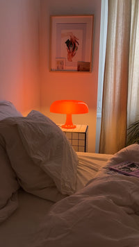 Gemütliches Schlafzimmer mit weißer Bettwäsche und leuchtender orangefarbener Midcentury-Tischlampe auf weißem Fliesentisch. An der Wand ein gerahmtes Kunstbild, beiger Vorhang am Fenster und Magazine auf dem Bett.