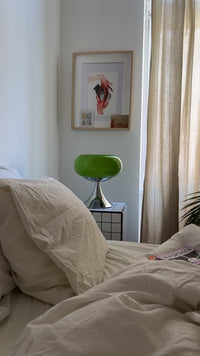 Die grüne Mid-Century-Lampe auf einem karierten Beistelltisch neben einem Bett mit beiger Bettwäsche. Eine Zeitschrift liegt auf der Decke, sanftes Tageslicht fällt durch helle Vorhänge und verleiht dem Raum eine ruhige Atmosphäre.