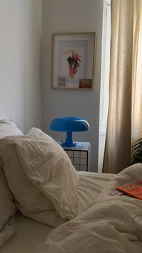 Die ikonische blaue Mushroom Lampe steht auf einem schwarz-weiß karierten Beistelltisch neben einem weißen Bett mit zerwühlter Decke. Wandbilder und eine helle Vorhangkulisse runden das moderne Interieur ab.