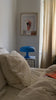 Die ikonische blaue Mushroom Lampe steht auf einem schwarz-weiß karierten Beistelltisch neben einem weißen Bett mit zerwühlter Decke. Wandbilder und eine helle Vorhangkulisse runden das moderne Interieur ab.