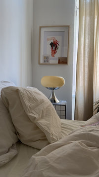 Cremefarbene Space Age Tischlampe auf einem karierten Nachttisch neben einem Bett mit beiger Bettwäsche. Sanfte, warme Farbtöne dominieren den Raum. An der Wand hängen Collagen und Kunstprints, die den gemütlichen Charakter unterstreichen.