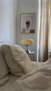 Cremefarbene Space Age Tischlampe auf einem karierten Nachttisch neben einem Bett mit beiger Bettwäsche. Sanfte, warme Farbtöne dominieren den Raum. An der Wand hängen Collagen und Kunstprints, die den gemütlichen Charakter unterstreichen.