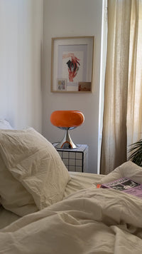 Die orangefarbene Space Age Tischlampe steht auf einem karierten Beistelltisch neben einem Bett mit weißer Bettwäsche. Die sanfte Beleuchtung schafft eine beruhigende Atmosphäre. Eine Zeitschrift liegt locker auf der Decke.