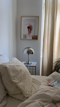 Helles Schlafzimmer mit beigem Bettzeug und verchromter Midcentury-Tischlampe auf weißem Fliesentisch. An der Wand ein gerahmtes Kunstbild mit roter Hand, daneben beigefarbener Vorhang und Zeitschriften auf dem Bett.