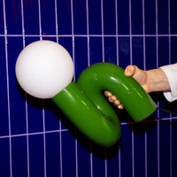 Snake Lampe Grün im organischen Design mit weißer Glaskugel vor blauem Fliesenspiegel gehalten.